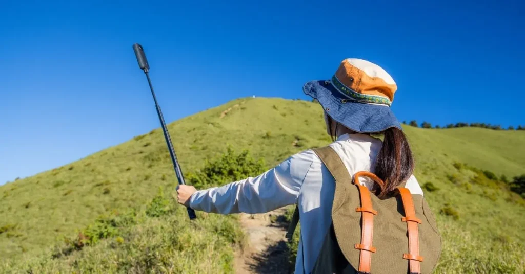 A woman hiking on a grassy hill with a trekking pole and wearing a backpack and a wide-brimmed hat on a sunny day.