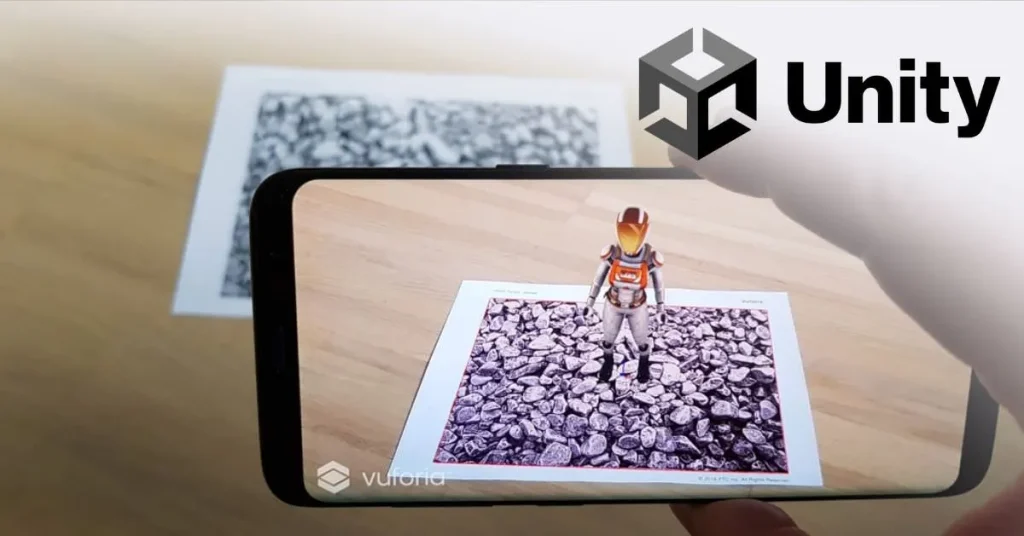 A smartphone displaying an augmented reality image of a 3D astronaut standing on a rocky surface, with a Unity logo in the background and a printed image marker on the table.