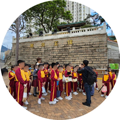 A group of students in uniform are gathered outdoors in front of a stone wall, with one person appearing to take a photo of them.